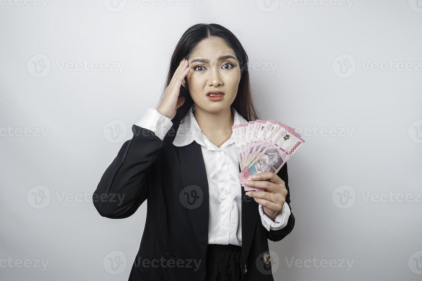 eine verwirrte junge geschäftsfrau trägt einen schwarzen anzug und hält bargeld in indonesischer rupiah, das durch weißen hintergrund isoliert wird foto