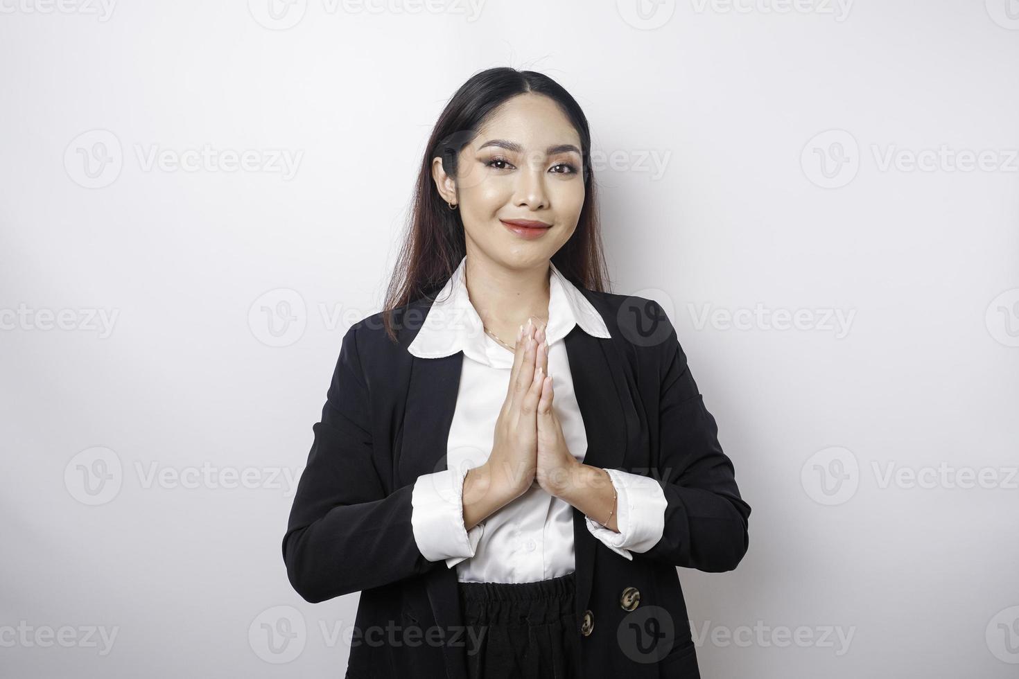 eine lächelnde junge asiatische mitarbeiterin, die einen schwarzen anzug trägt, gestikuliert einen traditionellen gruß, der über weißem hintergrund isoliert wird foto