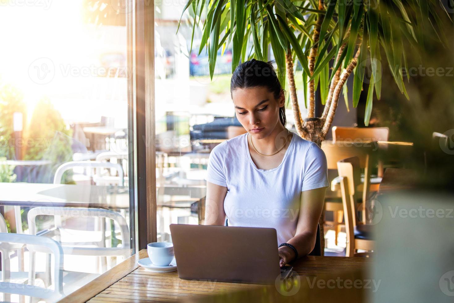 junges schönes Mädchen, das an einem Tisch in einem Café am Fenster sitzt und Cappuccino-Kaffee trinkt. schöne frau, die mit laptop vom café arbeitet. attraktive Frau, die in einem Café mit einem Laptop sitzt foto