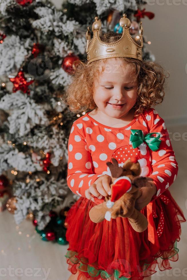 kleines lockiges mädchen in einem karnevalskleid versteckte ihr gesicht hinter glänzenden weihnachtsbaumspielzeugsternen. kind in einem roten kleid mit einem weihnachtsmanndruck auf dem hintergrund eines weihnachtsbaums. hochwertiges Foto
