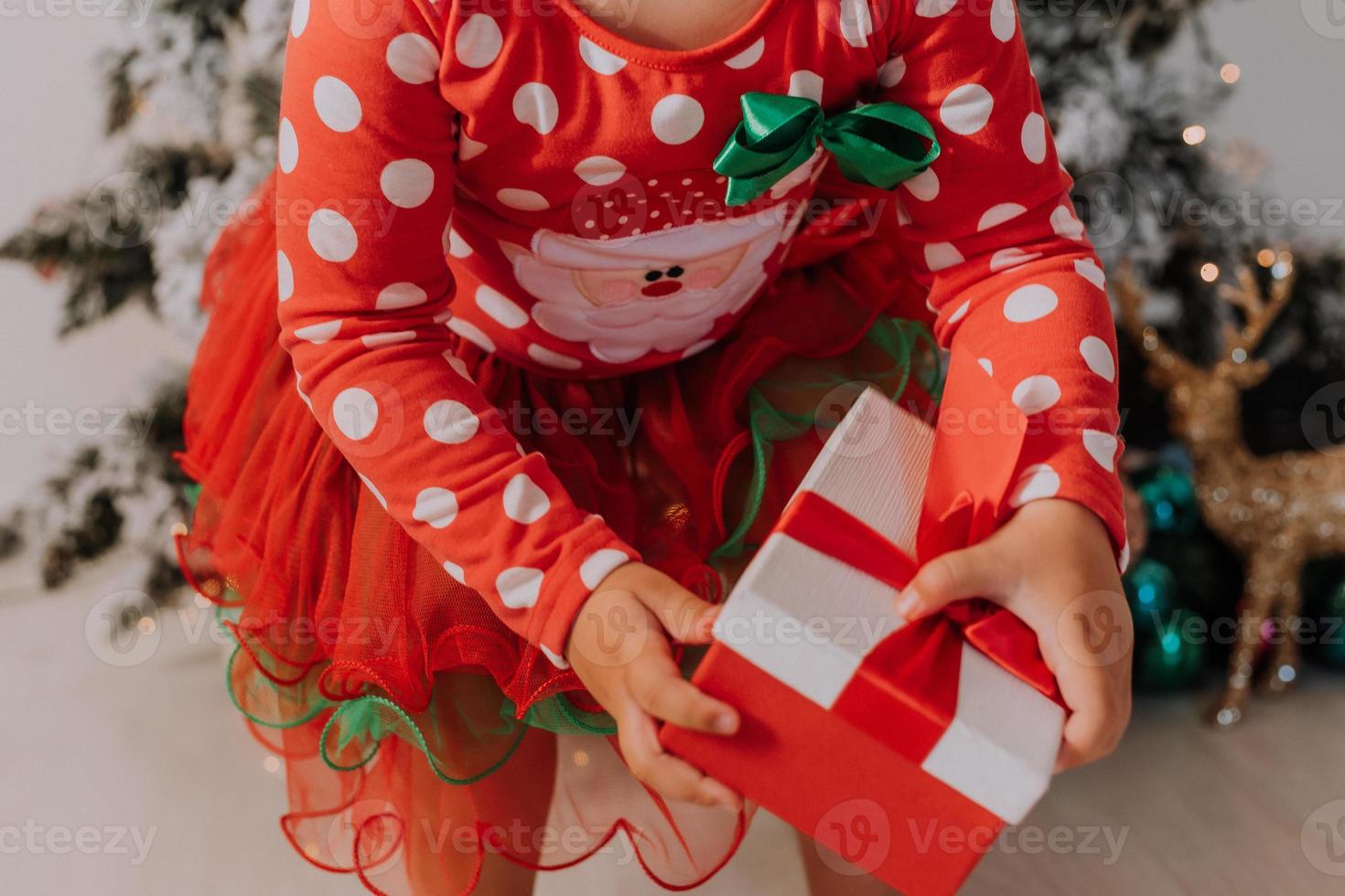 kleines lockiges mädchen in einem karnevalskleid versteckte ihr gesicht hinter glänzenden weihnachtsbaumspielzeugsternen. kind in einem roten kleid mit einem weihnachtsmanndruck auf dem hintergrund eines weihnachtsbaums. hochwertiges Foto