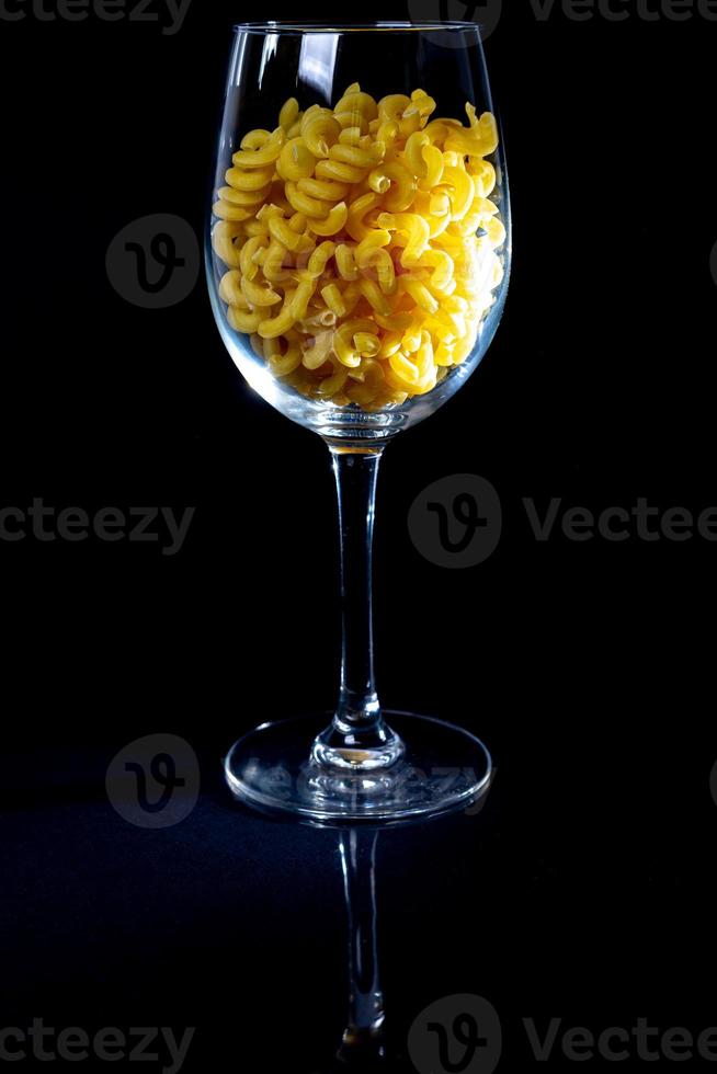 rohe Nudeln im Glas, Weinglas. im Eimer. rohe Nudeln auf schwarzem Hintergrund. Vorderansicht rohe Nudeln, aus der Hand gefallen, Platz für Text, gelbe lange Spaghettistrohhalme, foto