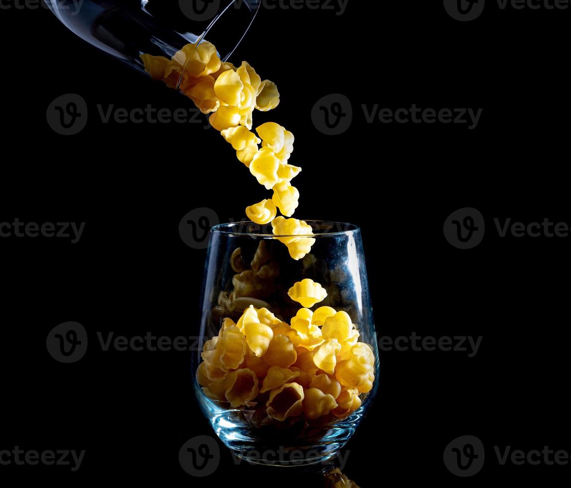 rohe Nudeln im Glas, Weinglas. im Eimer. rohe Nudeln auf schwarzem Hintergrund. Vorderansicht rohe Nudeln, aus der Hand gefallen, Platz für Text, gelbe lange Spaghettistrohhalme, foto