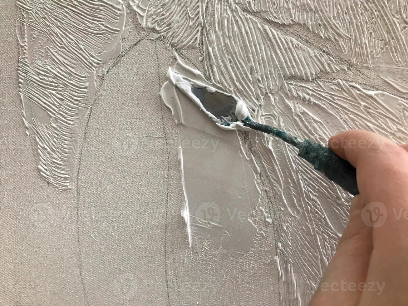 Die Hand des Künstlers verputzt die Leinwand mit einem kleinen Spachtel mit volumetrischer weißer Acrylfarbe in der kreativen Kunstwerkstatt des Künstlers mit Farben und Pinseln auf dem Tisch der Kunstschule foto
