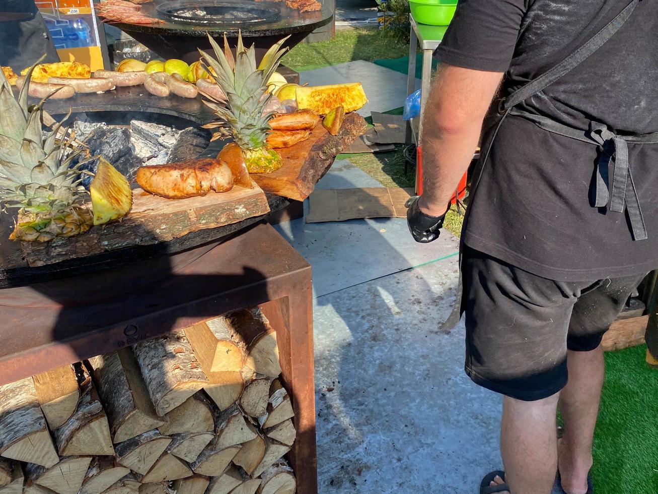 Leute grillen Würstchen, Fleisch und Ananas auf einem Rost über einem offenen Feuer mit Holz und Ananas unter freiem Himmel bei einem Musikfestival foto