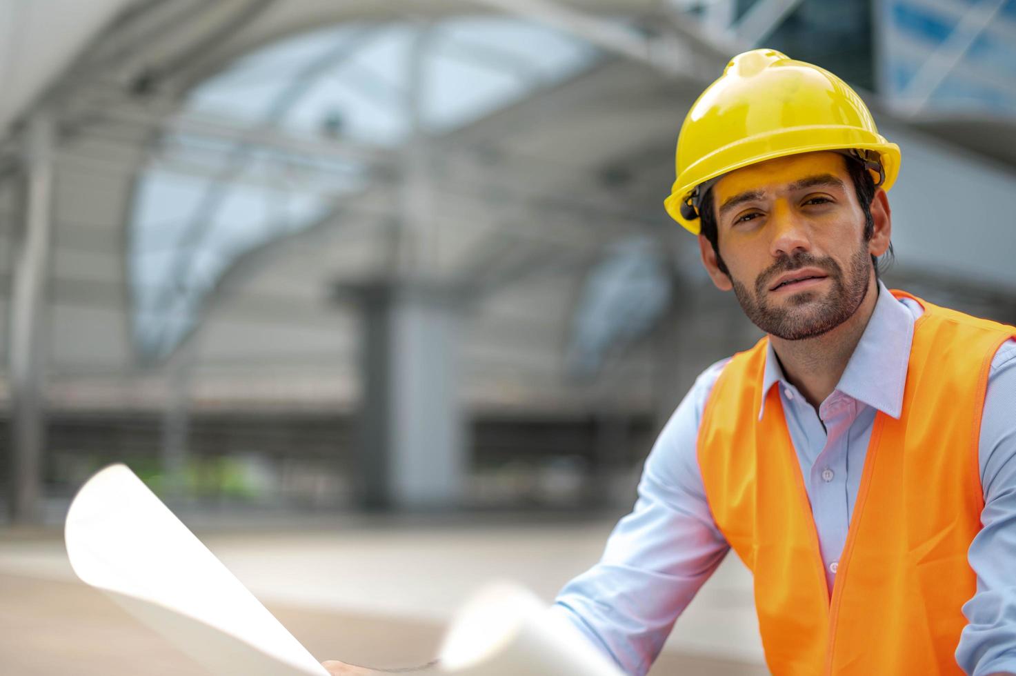 kaukasischer ingenieur, der eine orangefarbene weste und einen großen harten hut trägt und andererseits den weißen grundriss in der vorortarbeit der stadtmitte hält. foto