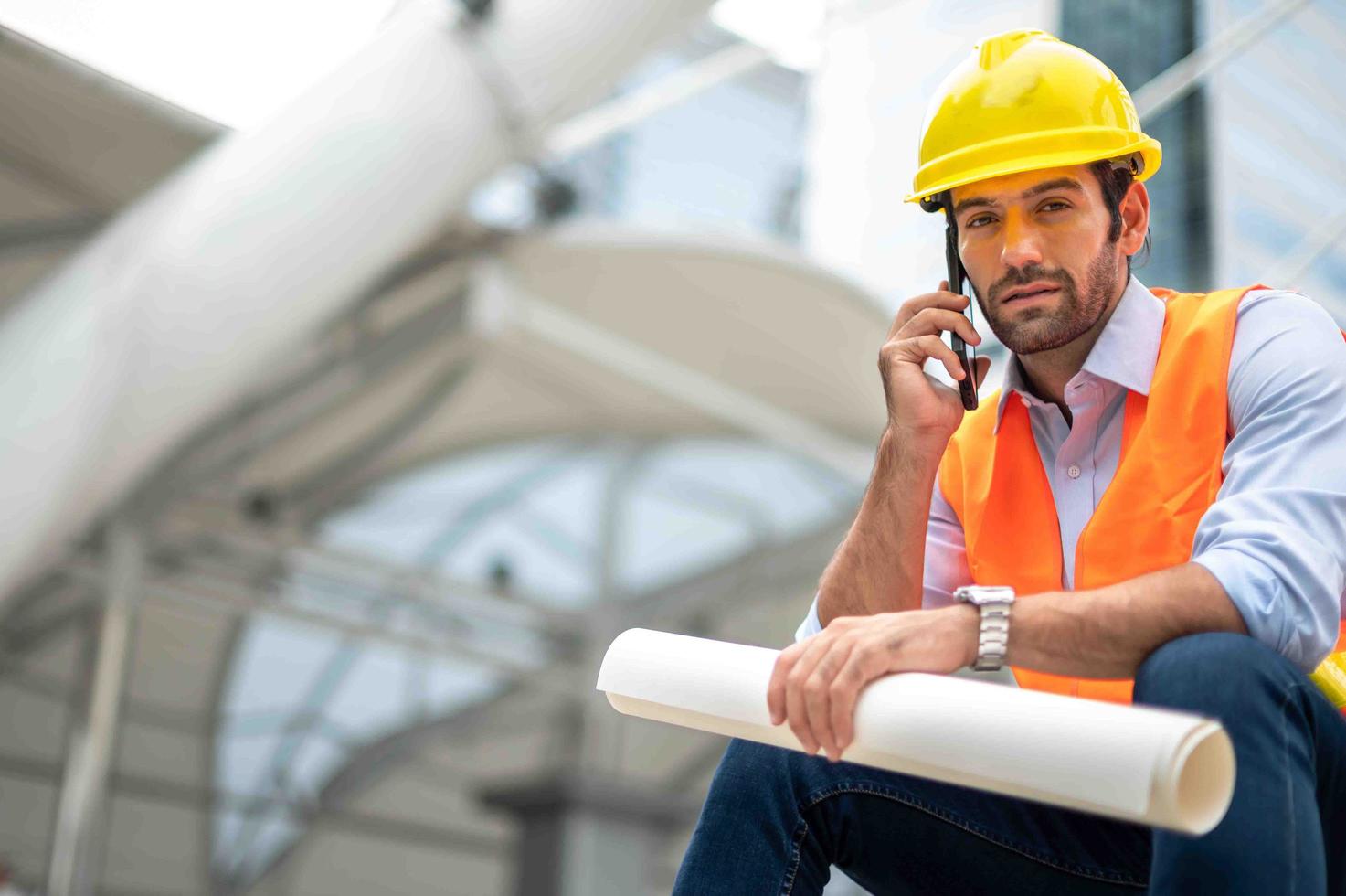 Kaukasischer Ingenieur verwendet ein Smartphone zum Reden, trägt eine orangefarbene Weste und einen großen Schutzhelm und hält andererseits den weißen Grundriss in der Baustelle der Innenstadt. foto