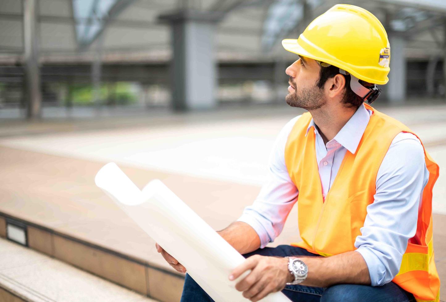 kaukasischer ingenieur, der eine orangefarbene weste und einen großen harten hut trägt und andererseits den weißen grundriss in der vorortarbeit der stadtmitte hält. foto