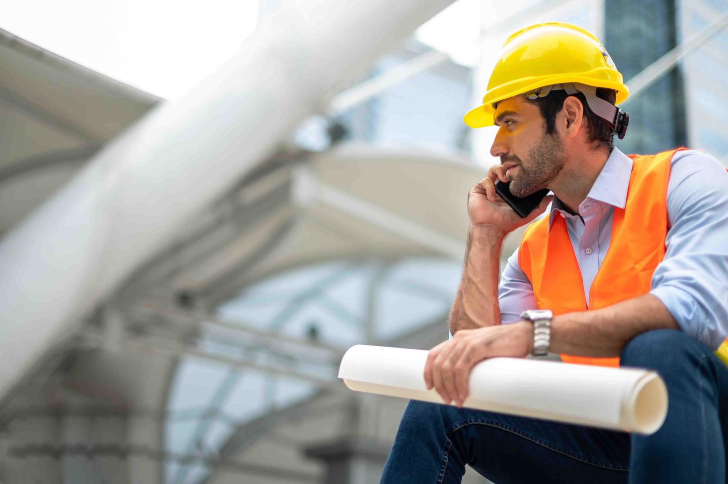 Kaukasischer Ingenieur verwendet ein Smartphone zum Reden, trägt eine orangefarbene Weste und einen großen Schutzhelm und hält andererseits den weißen Grundriss in der Baustelle der Innenstadt. foto