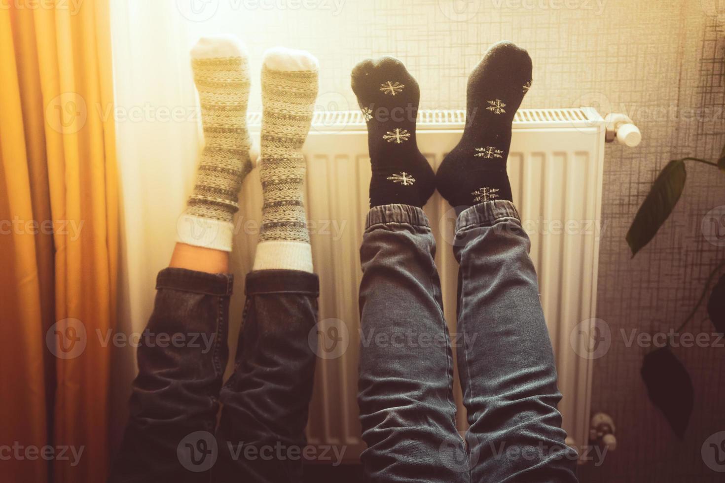 Paar hält die Beine hoch und wärmt im Winter die Füße in einem kalten Zuhause auf dem Heizkörper mit gemütlichen winterlichen Wollsocken. valentinstag lustig zusammen warme füße am heizkörper am fenster foto