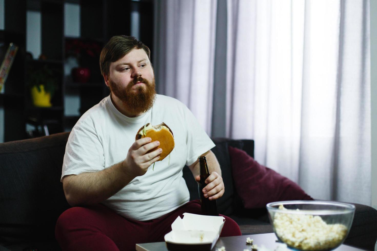 dicker Mann isst Burger mit Bier, das am Tisch vor dem Fernsehen sitzt foto