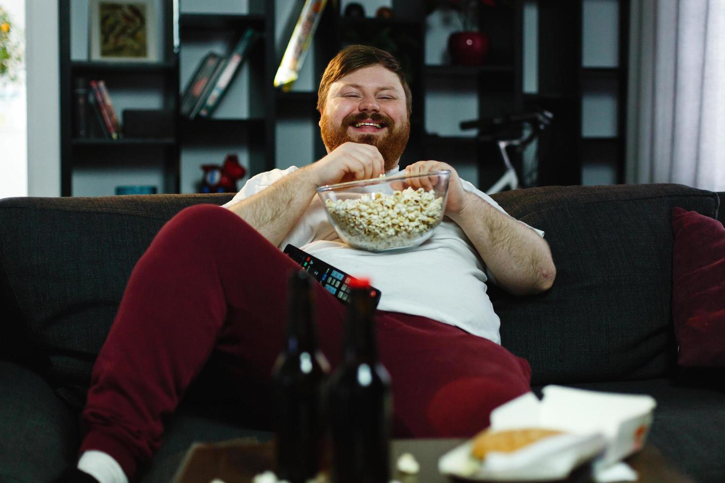 lachender dicker Mann sitzt auf dem Sofa, isst Popcorn und sieht fern foto