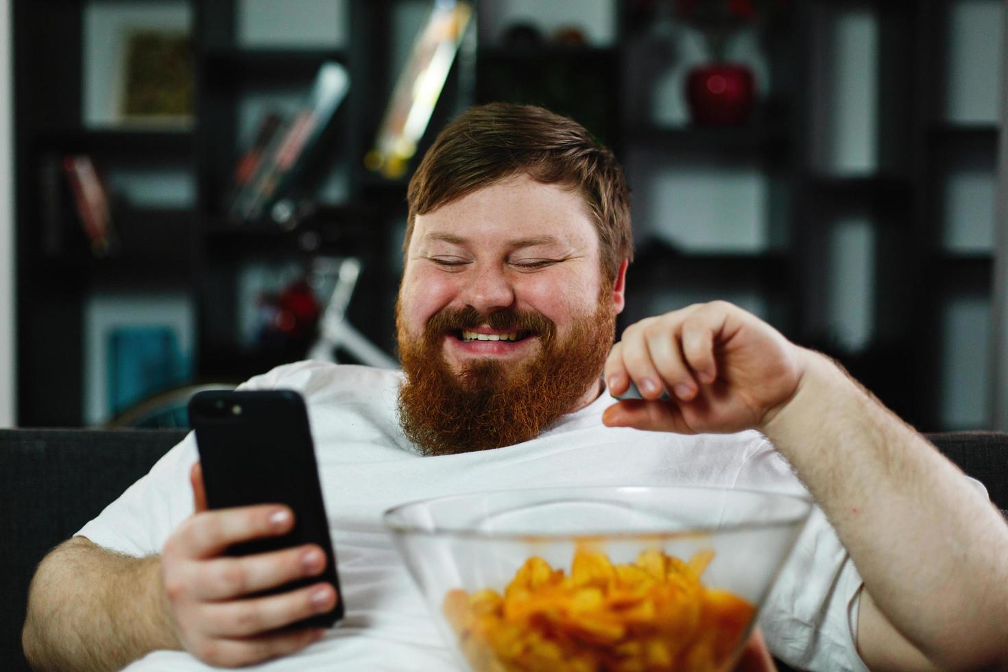 Mann mit einem Bier, das Chips isst und auf sein Telefon schaut foto