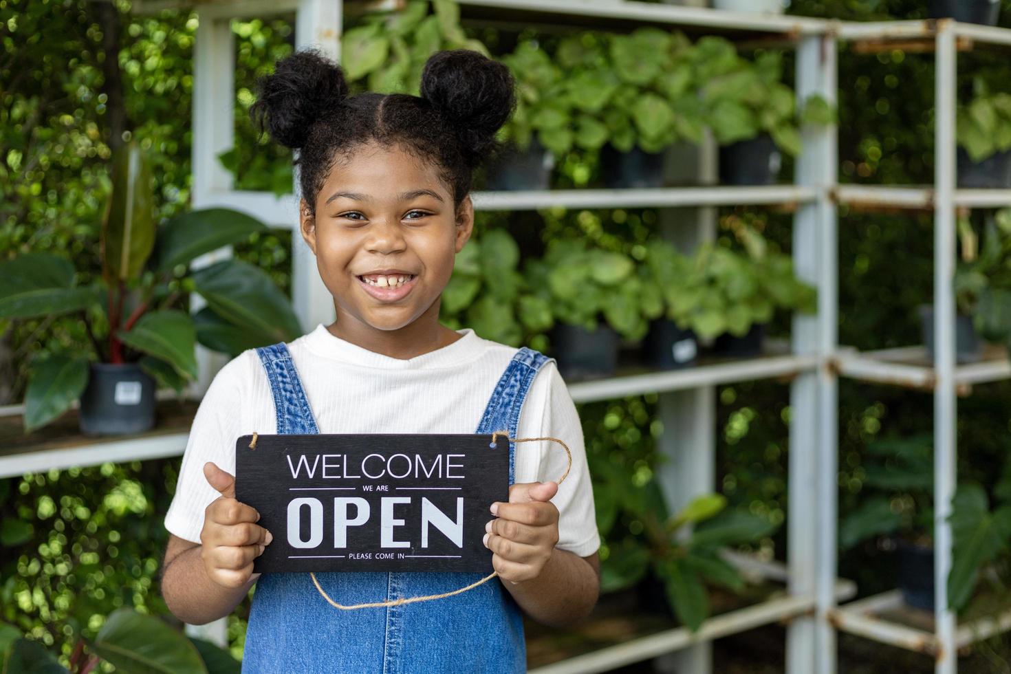 Porträt eines afroamerikanischen Mädchens, das ein Willkommensschild für das Gartenzentrum ihrer Familie für Baumschulen und Zimmerpflanzengeschäft hält foto