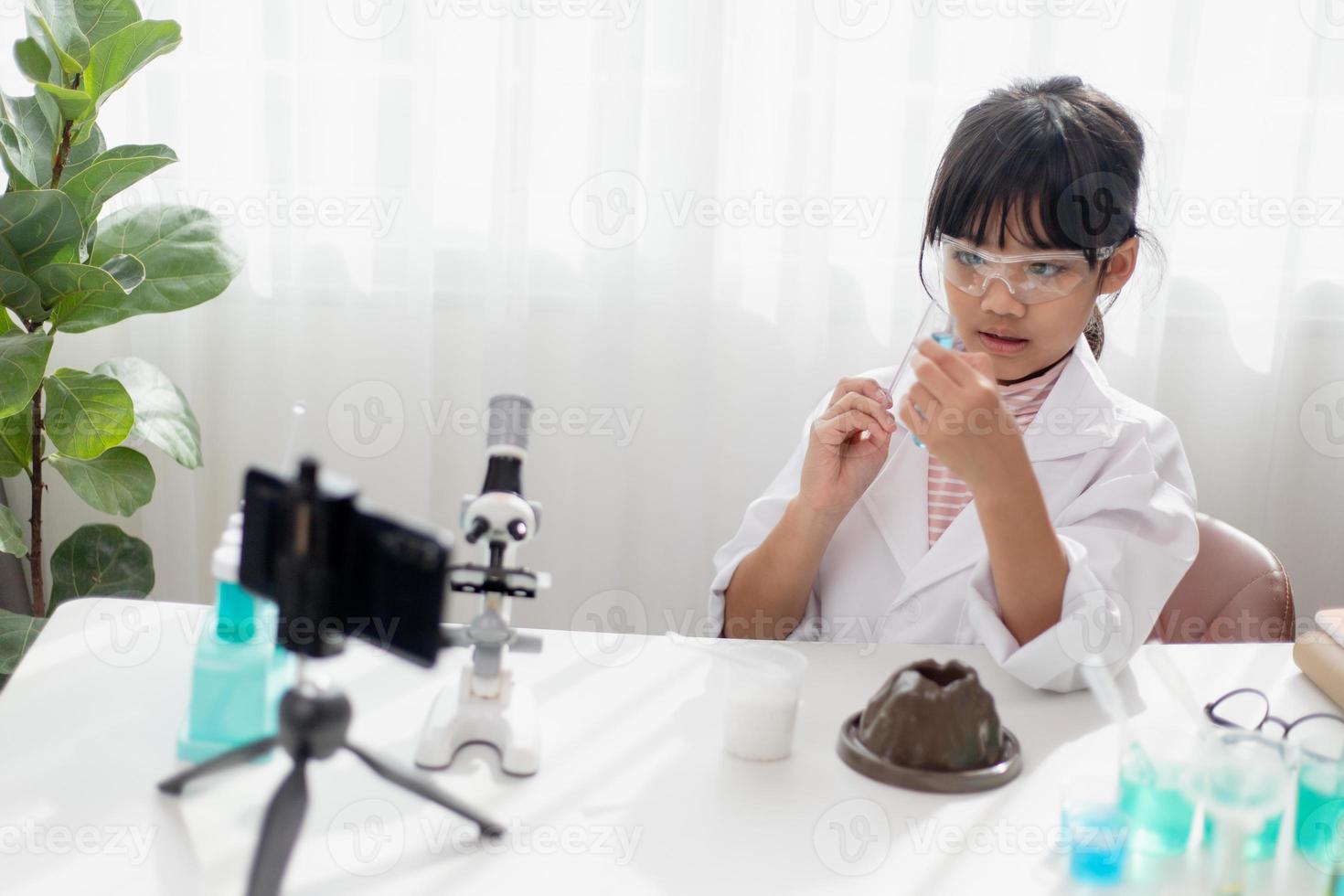 asiatisches schulmädchen, das einfache chemieexperimente macht und ein video für seine anhänger aufnimmt, junges bloggerkind, das vor der kamera für vlog posiert, kinder machen vlog für social-media-kanalkonzept. foto