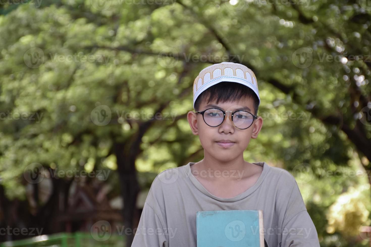 Der junge asiatische muslimische Junge trägt eine Brille, sitzt im Schulpark und liest sein Buch in seiner Freizeit, bevor er nach Hause geht, weicher und selektiver Fokus. foto