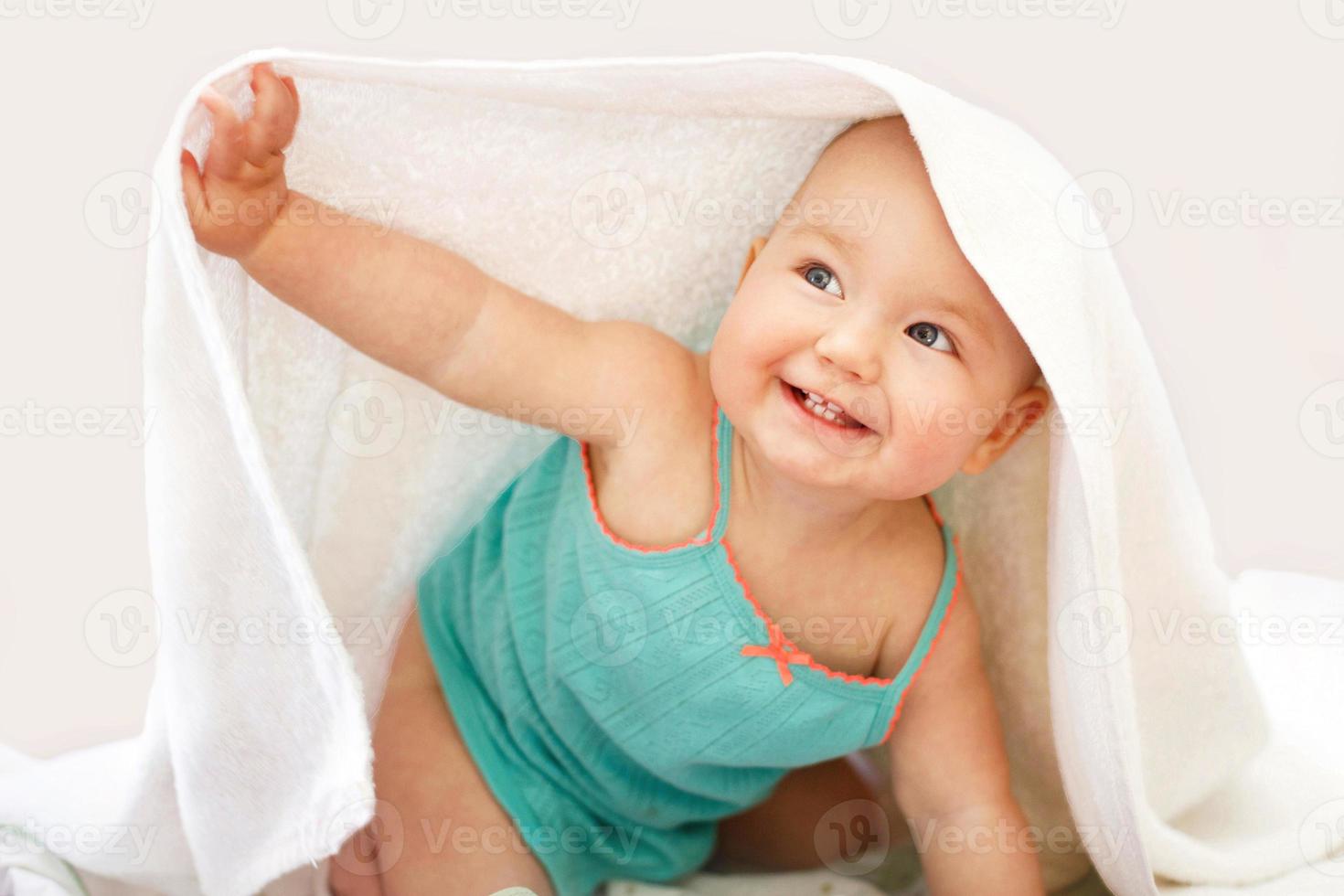 Süßes lächelndes Baby, das unter einem weißen Handtuch auf weißem Hintergrund in die Kamera schaut. Porträt eines süßen Kindes foto
