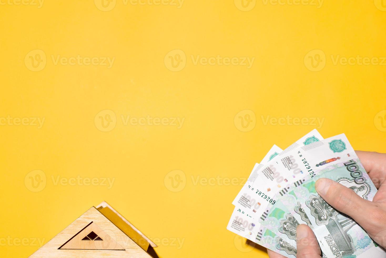 Modell des Holzdaches des Hauses und der Hände des Mannes, die Geld halten, Draufsicht. das konzept des kaufs von immobilien, stromrechnungen, versicherungen, investitionen. gelber hintergrund, kopierraum foto