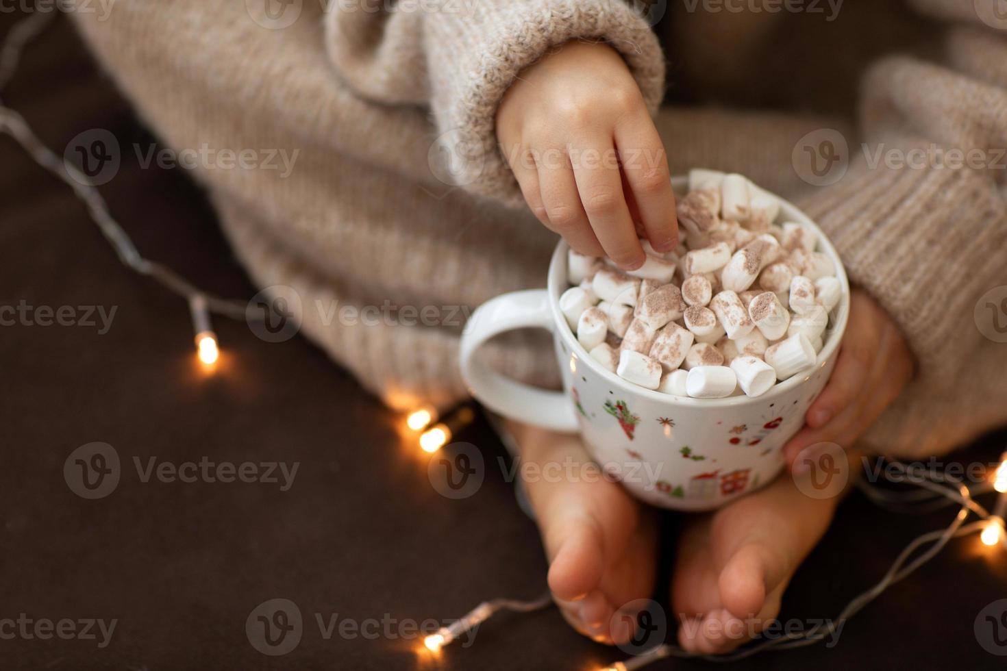 barfuß und hände des kindes halten tasse heißen kakao marshmallow nahaufnahme girlandenlichter. flauschiger warmer beige gestrickter pullover. weihnachtskonzept, urlaub. frohes neues jahr. kindermädchen, das innen auf sofa sitzt foto
