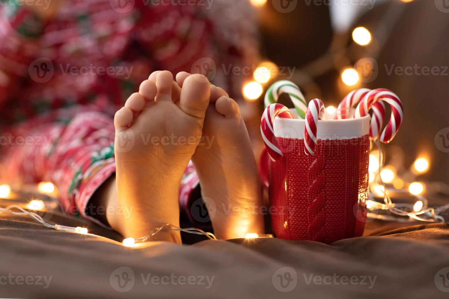 Nahaufnahme Kind nackte Füße Kind im roten Weihnachtspyjama halten Becher Tasse gestreift grün rot Zuckerstangen bokeh Lichter Hintergrund. Neujahr foto