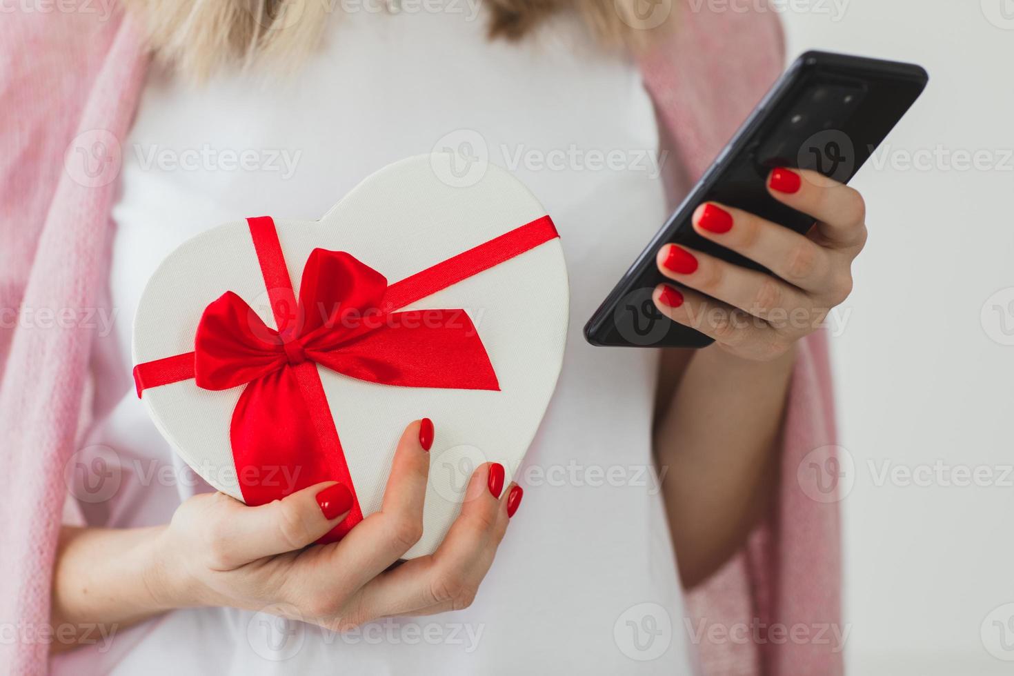 Nahaufnahme der Hände einer Frau mit roter Maniküre hält Geschenkbox Herz und Smartphone. Online-Lieferservice für Geschenke, Weihnachtseinkäufe, Kompliment. Valentinstag foto