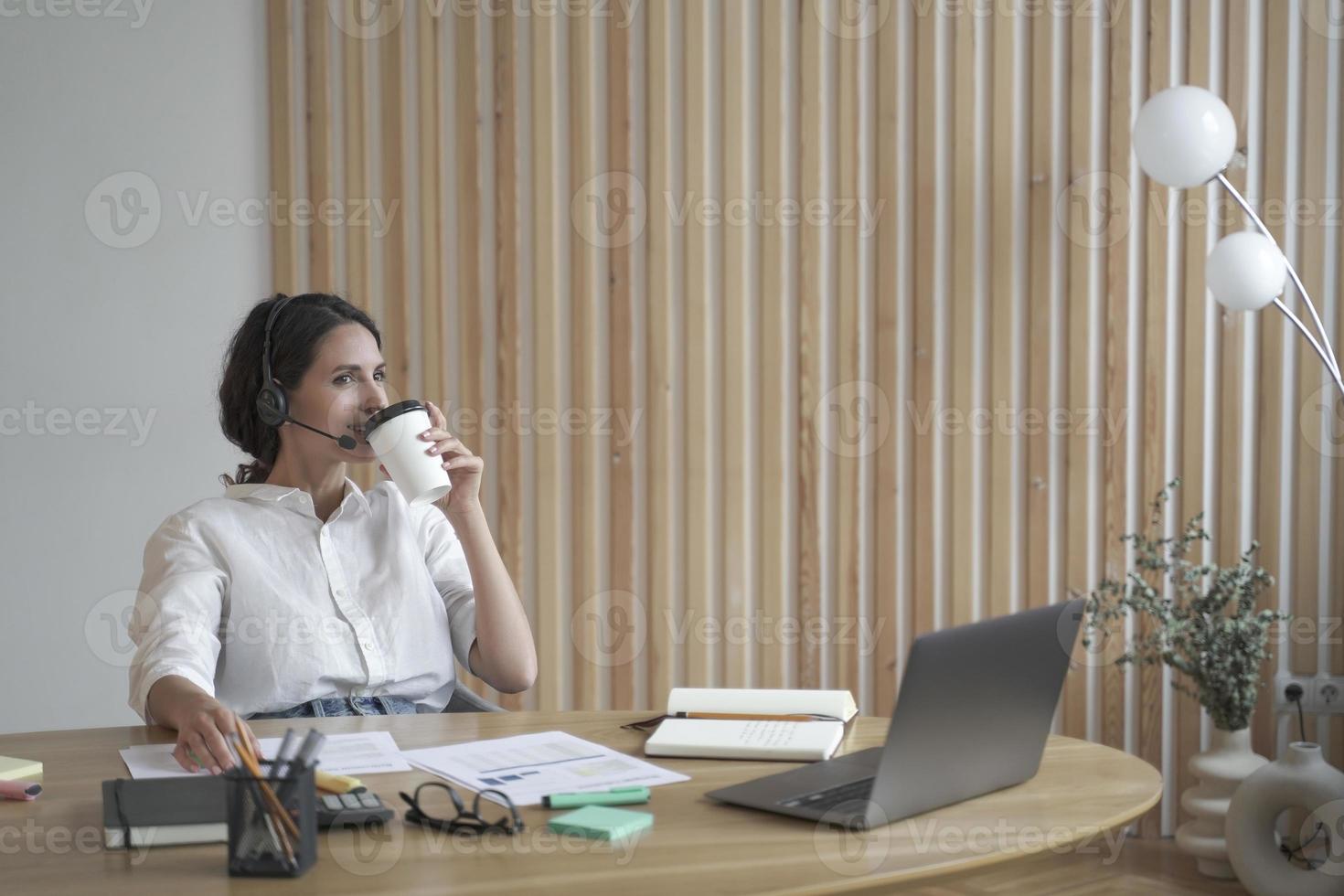 Zufriedene positive spanische Unternehmerin im drahtlosen Kopfhörer, die Kaffee im Büro trinkt foto