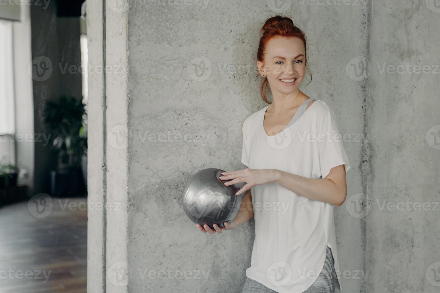 Rothaarige positive Frau, die vor dem Pilates-Kurs mit Fitball in der Hand an einer grauen Wand steht foto