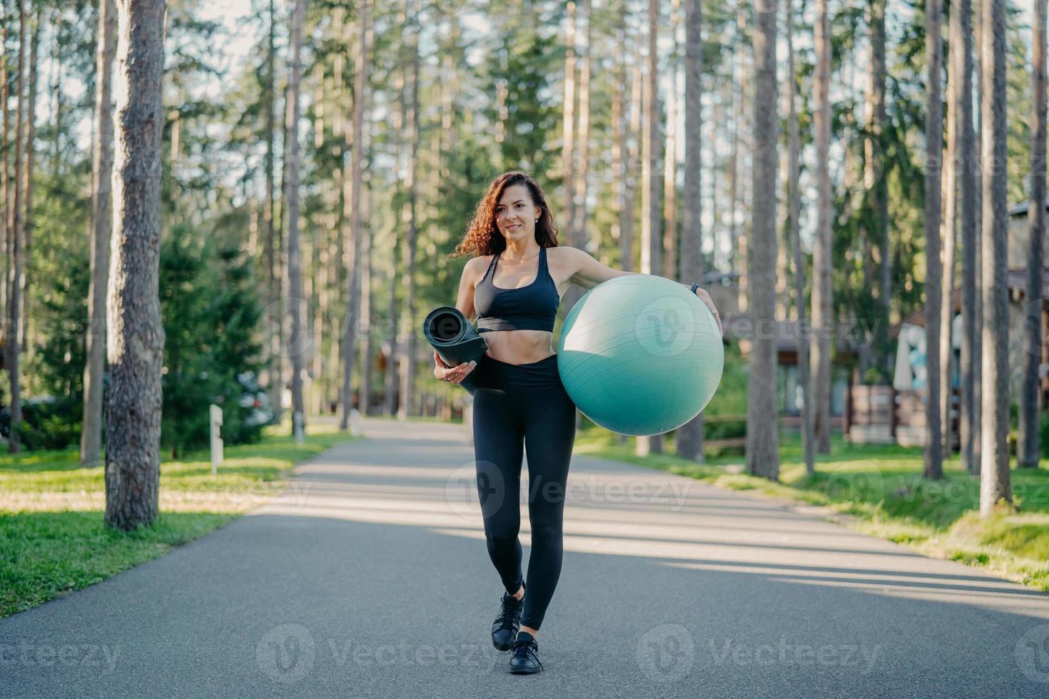 horizontale aufnahme einer aktiven schlanken frau in sportbekleidung, trägt fitball und aufgerollte karemat, geht auf der straße in der nähe von bäumen spazieren, wird trainieren, atmet frische luft, führt einen gesunden sportlichen lebensstil foto