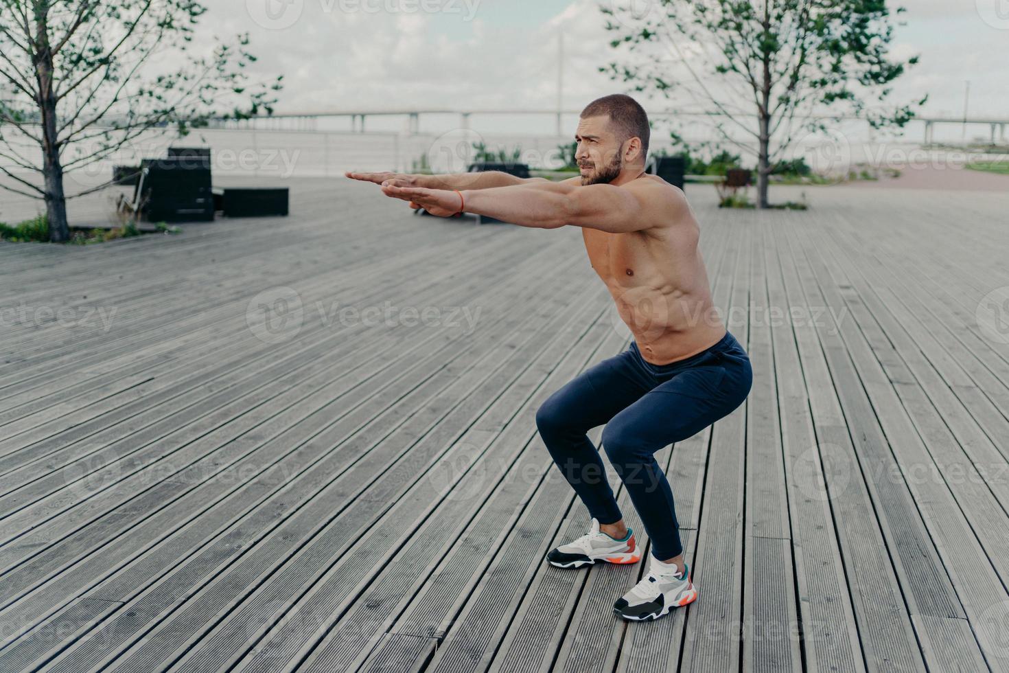 Ganzkörperaufnahme eines aktiven Mannes macht Hockübungen mit nacktem Oberkörper und hat muskulöse Körperhaltungen draußen. starker athletischer fitter mann in sportbekleidungsübungen im freien. selbstbewusster Fitnesstrainer foto