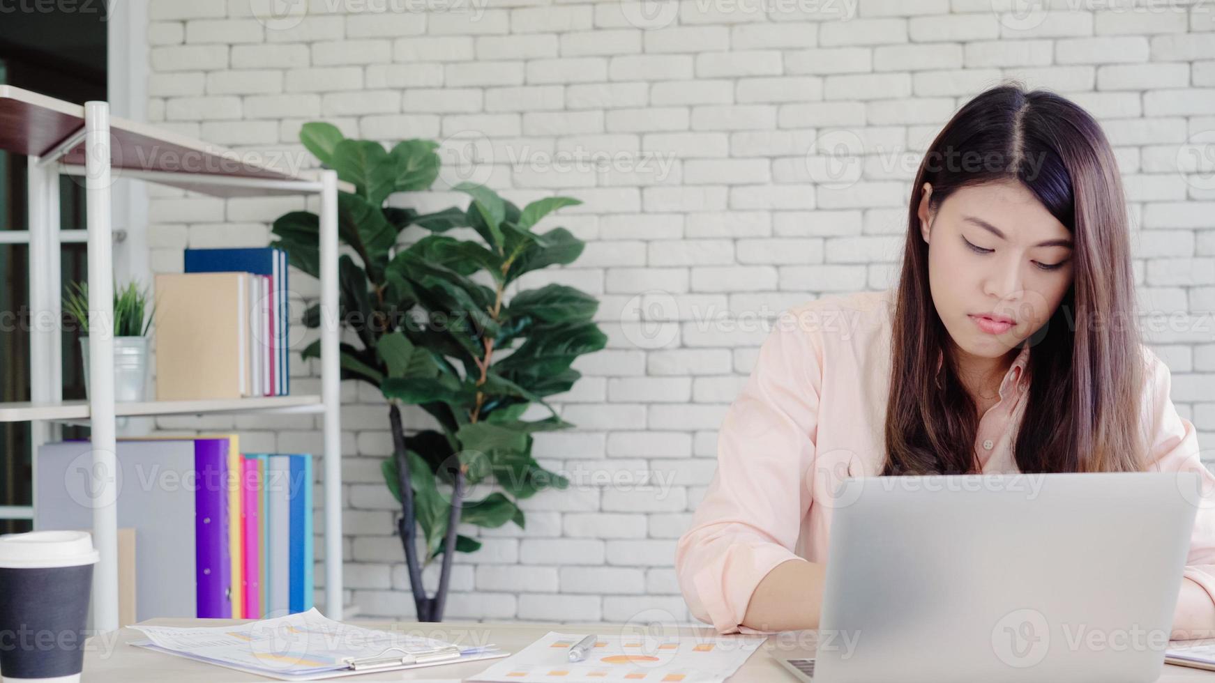 schöne junge lächelnde asiatische Frau, die Laptop auf Schreibtisch im Wohnzimmer zu Hause arbeitet. asiatische Geschäftsfrau, die Notizbuchdokumentfinanzierung und -rechner im Heimbüro schreibt. Zeit zu Hause Konzept genießen. foto