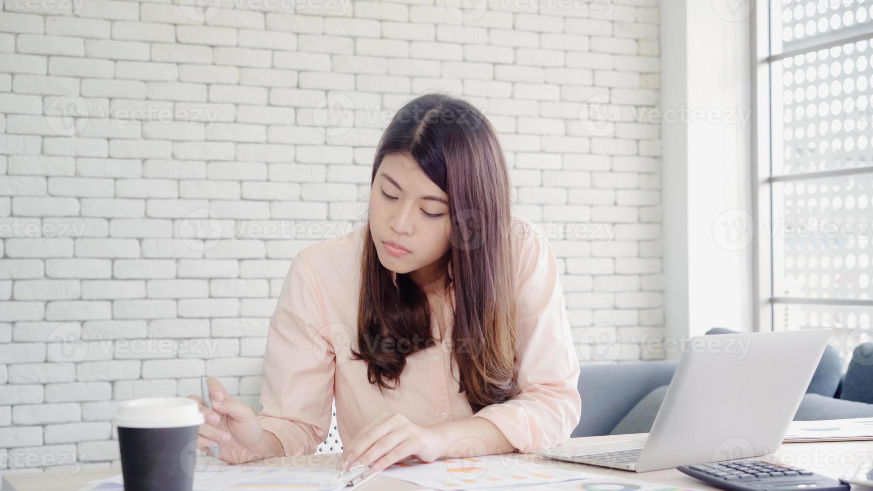 schöne junge lächelnde asiatische Frau, die Laptop auf Schreibtisch im Wohnzimmer zu Hause arbeitet. asiatische Geschäftsfrau, die Notizbuchdokumentfinanzierung und -rechner im Heimbüro schreibt. Zeit zu Hause Konzept genießen. foto