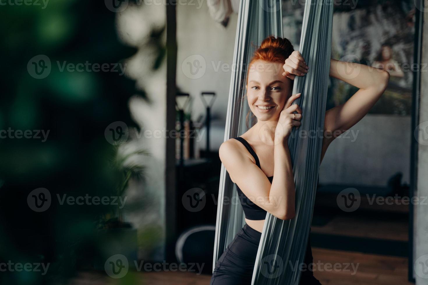 Junge rothaarige Frau, die in der Hängematte sitzt, während sie den Aerial Yoga-Kurs besucht foto