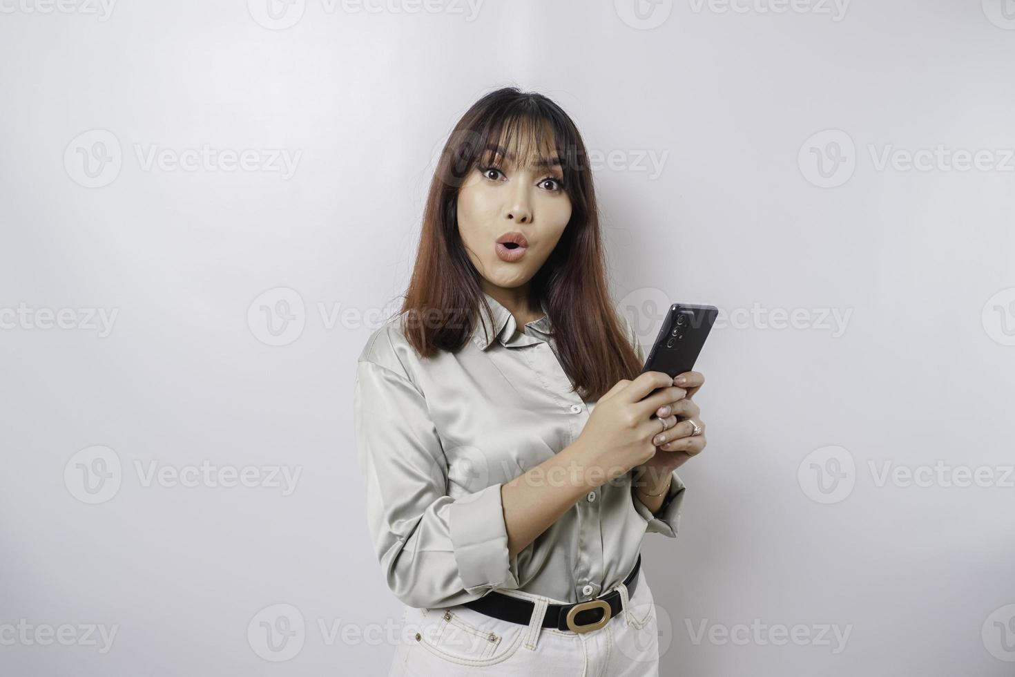 Schockierte asiatische Frau, die ein salbeigrünes Hemd trägt und ihr Telefon hält, isoliert durch weißen Hintergrund foto