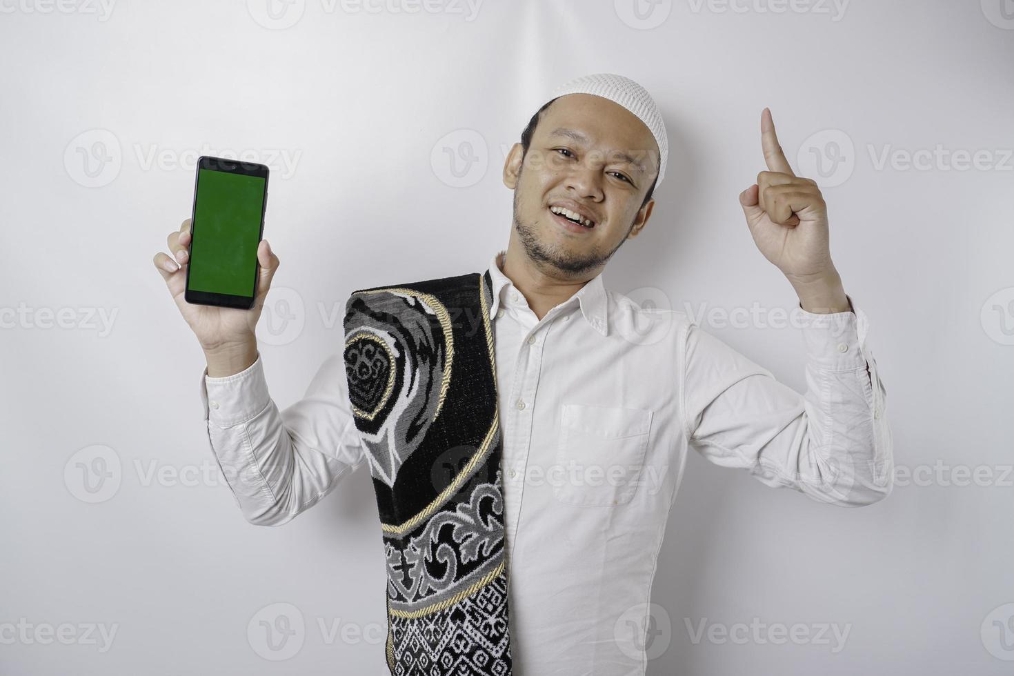 Ein Porträt eines glücklichen asiatischen muslimischen Mannes, der lächelt, während er auf seinem Telefon Platz für Kopien zeigt und nach oben zeigt, isoliert durch weißen Hintergrund foto