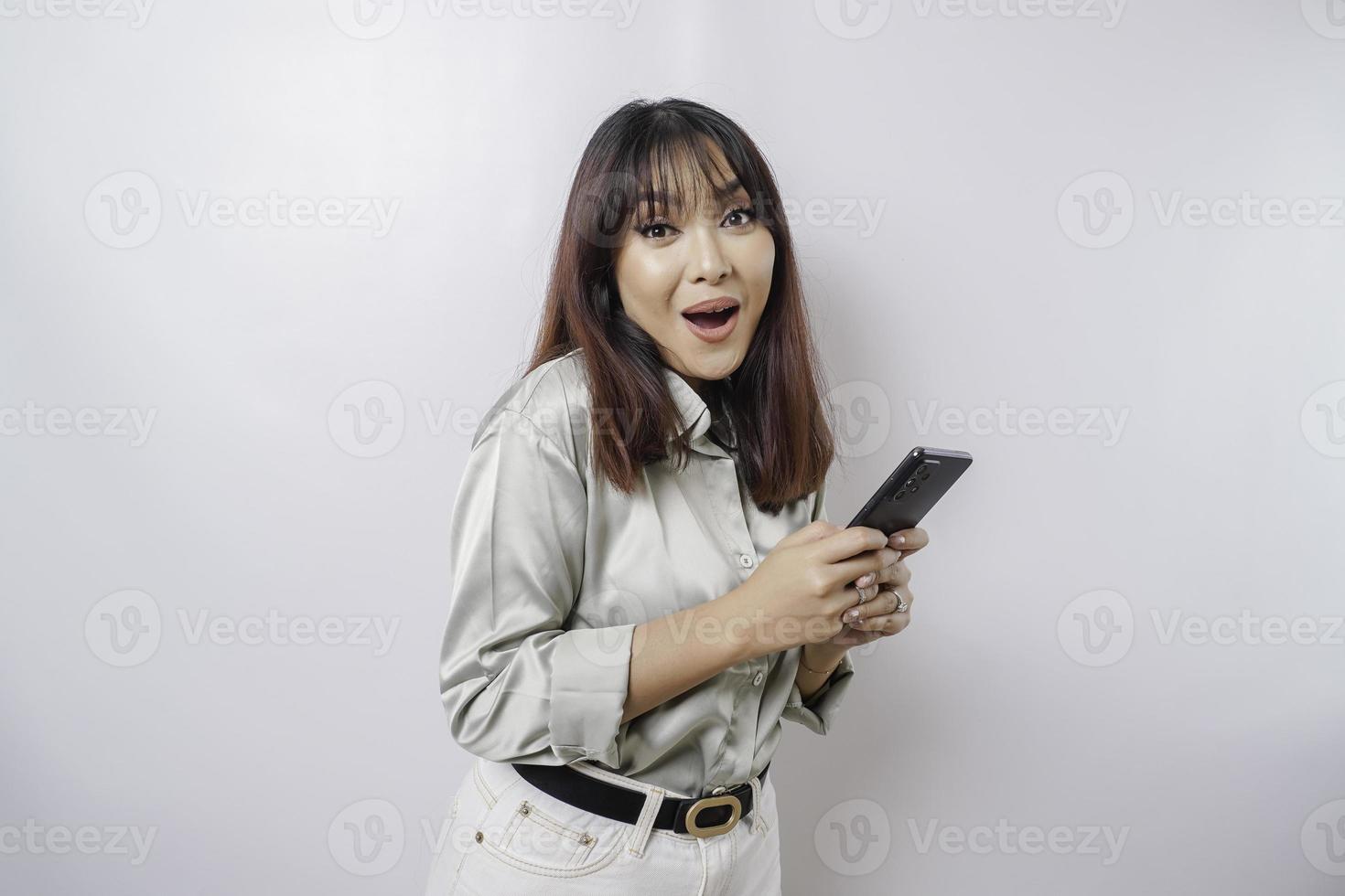 Schockierte asiatische Frau, die ein salbeigrünes Hemd trägt und ihr Telefon hält, isoliert durch weißen Hintergrund foto