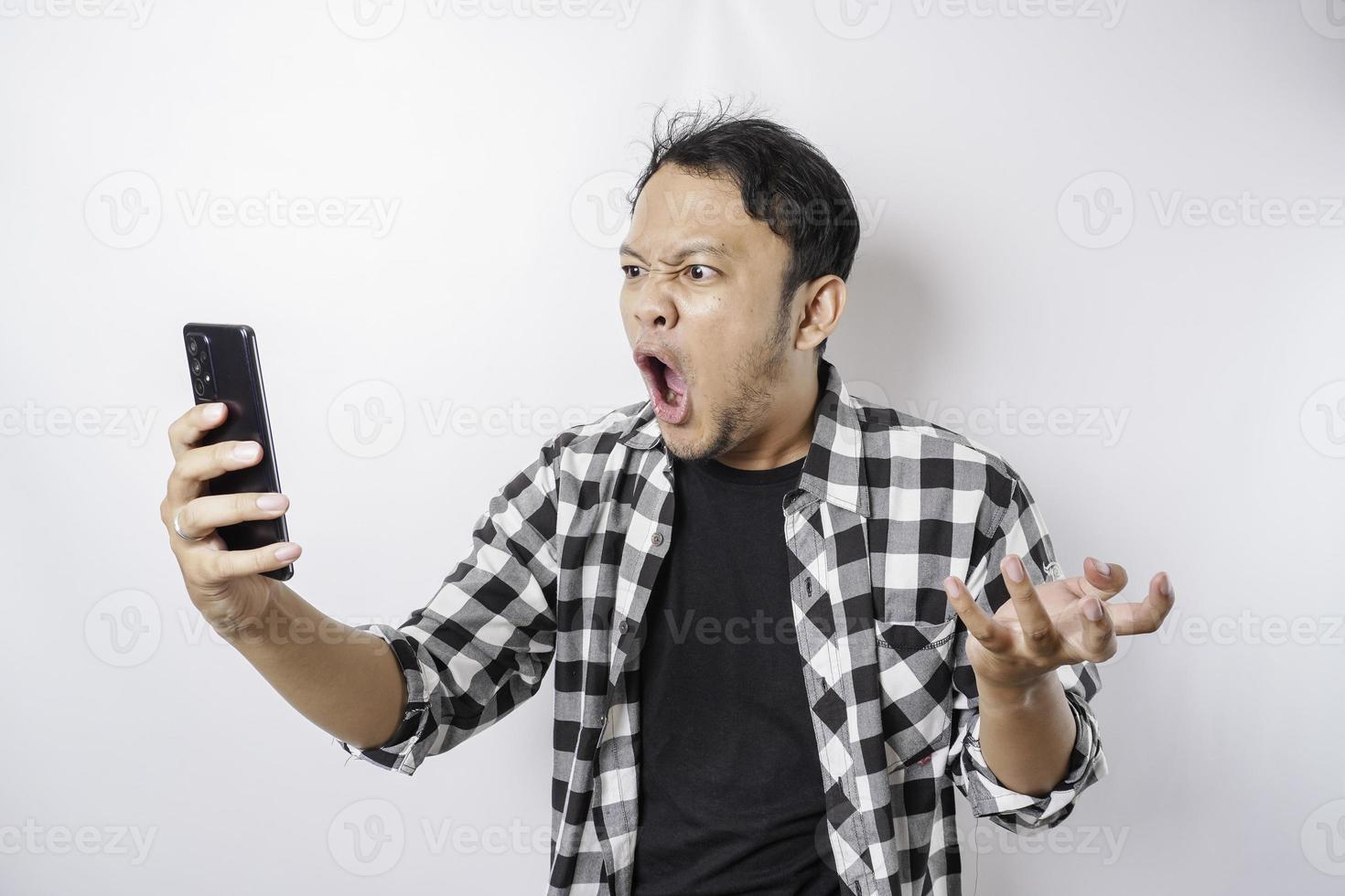 das wütende und wütende gesicht des asiatischen mannes im tartanhemd, während er sein telefon auf isoliertem weißem hintergrund hält. foto