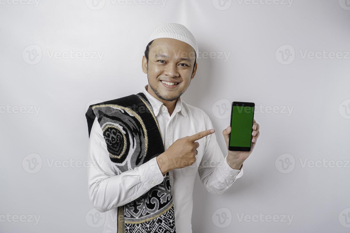 ein Porträt eines glücklichen asiatischen muslimischen Mannes, der lächelt, während er Kopienraum auf seinem Telefon zeigt, isoliert durch weißen Hintergrund foto