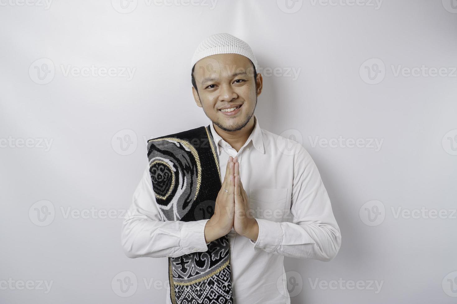 lächelnder junger asiatischer muslimischer Mann mit Gebetsteppich auf der Schulter, gestikulierender traditioneller Gruß einzeln auf weißem Hintergrund foto