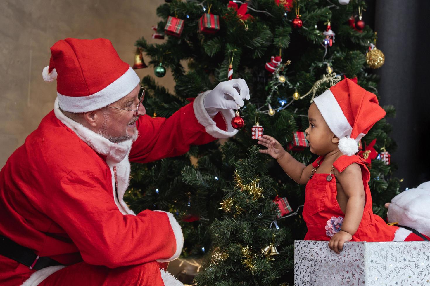 afroamerikanisches baby bekommt vom weihnachtsmann nachts eine zierkugel als geschenk am voll geschmückten weihnachtsbaum für die saisonfeier foto