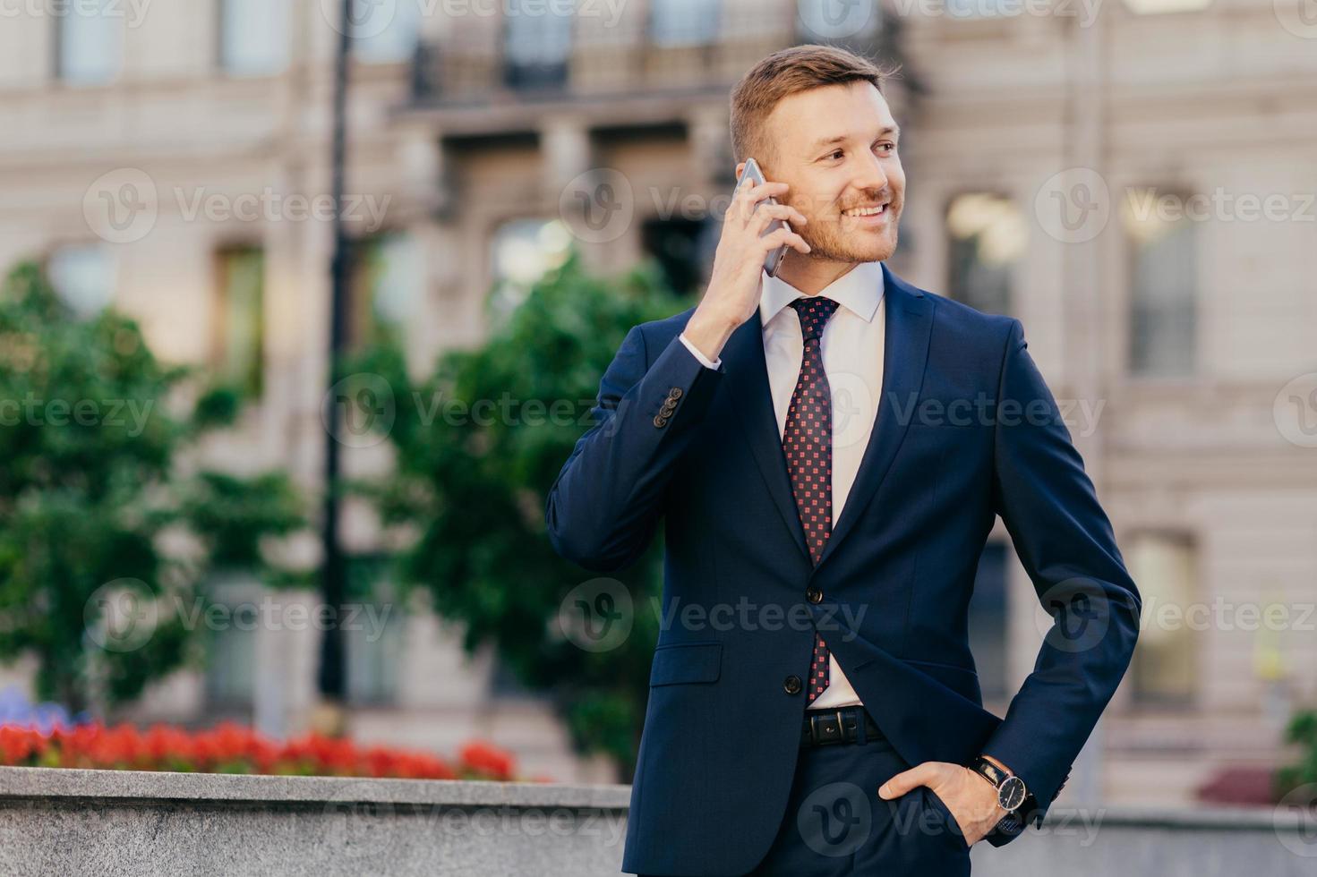 positiver erfolgreicher geschäftsmann teilt nachrichten mit freunden, freut sich über ein neues innovatives projekt, steht im freien, hält die hand in der tasche, ist formell gekleidet, bemerkt etwas in der ferne beiseite foto