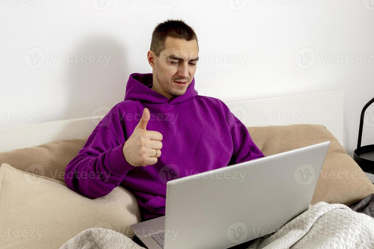 junger kaukasischer mann mit violettem kapuzenpulli, der auf bett sitzt und laptop-computer hält. mann, der notebook benutzt, um online zu lernen, freunde anrufen. Fernunterricht, Videoanruf. positives Vorzeichen. foto