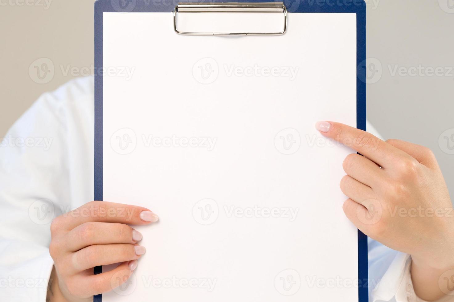 ein arzt in einem weißen kittel zeigt auf eine leere stelle auf einem papier. der arzt hält einen ordner mit einem leeren blatt in den händen. Platz für Kopien und Aufzeichnungen. medizinisches konzept.kopierraum foto