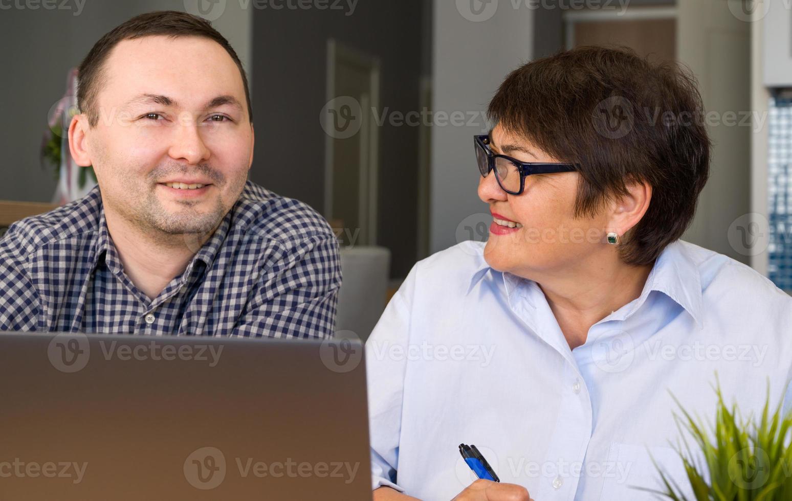 Seniorin schreibt Informationen auf, während ihr Sohn ihr erklärt, wie man die Anwendung auf dem Computer verwendet foto