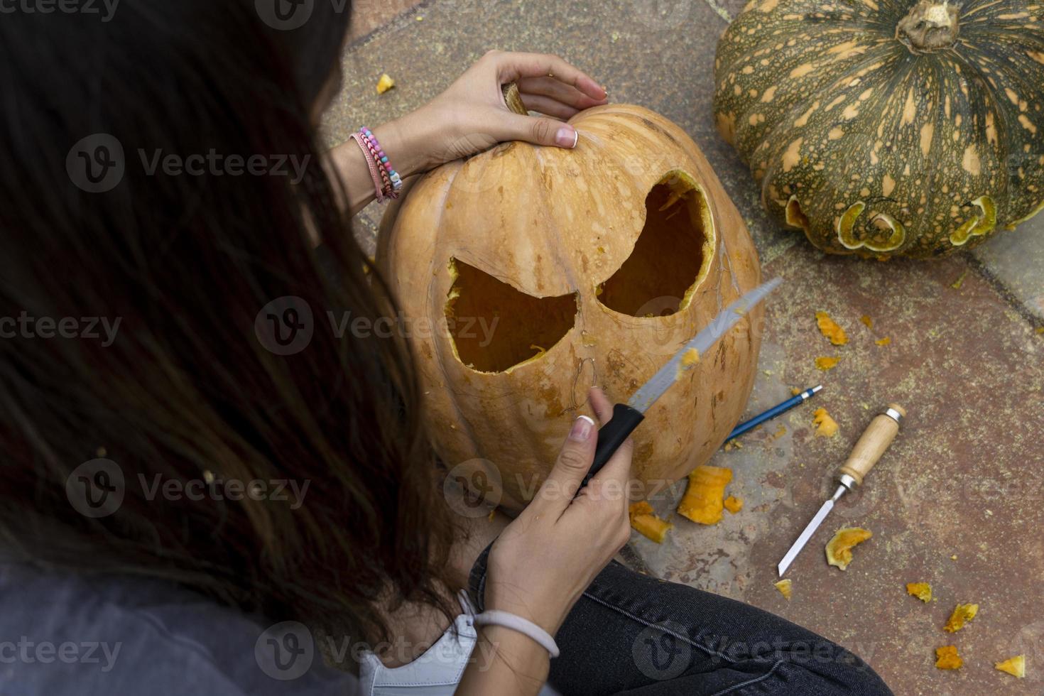 Frau, die großen orangefarbenen Kürbis für Halloween schnitzt, während sie zu Hause am Holztisch sitzt foto