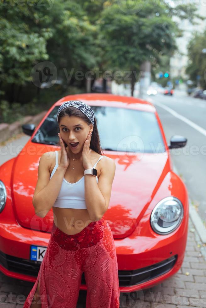 Porträt einer hübschen kaukasischen Frau, die gegen ein neues rotes Auto steht foto