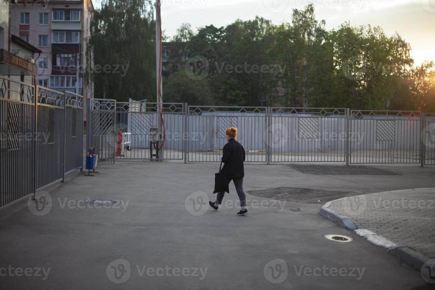 Ein Mädchen in Schwarz mit einer Tasche in der Hand geht die Straße hinunter zu den Häusern im Hintergrund. grauer Zaun, grüne Bäume. sonnenuntergang sonne, urbanes leben foto