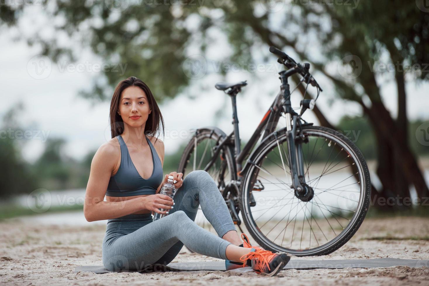 Vergessen Sie nicht, Wasser für Ihre Übungen mitzunehmen. Radfahrerin mit guter Körperform, die tagsüber neben ihrem Fahrrad am Strand sitzt foto