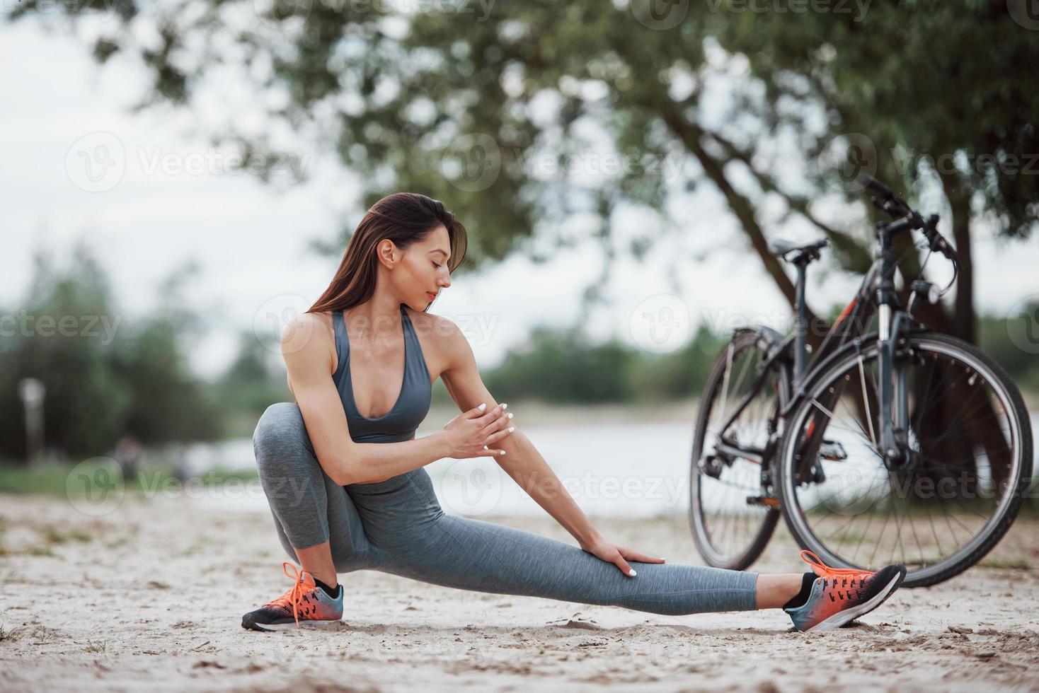 konzentrierter Blick. Radfahrerin mit guter Körperform, die tagsüber Yoga-Übungen macht und sich in der Nähe ihres Fahrrads am Strand dehnt foto