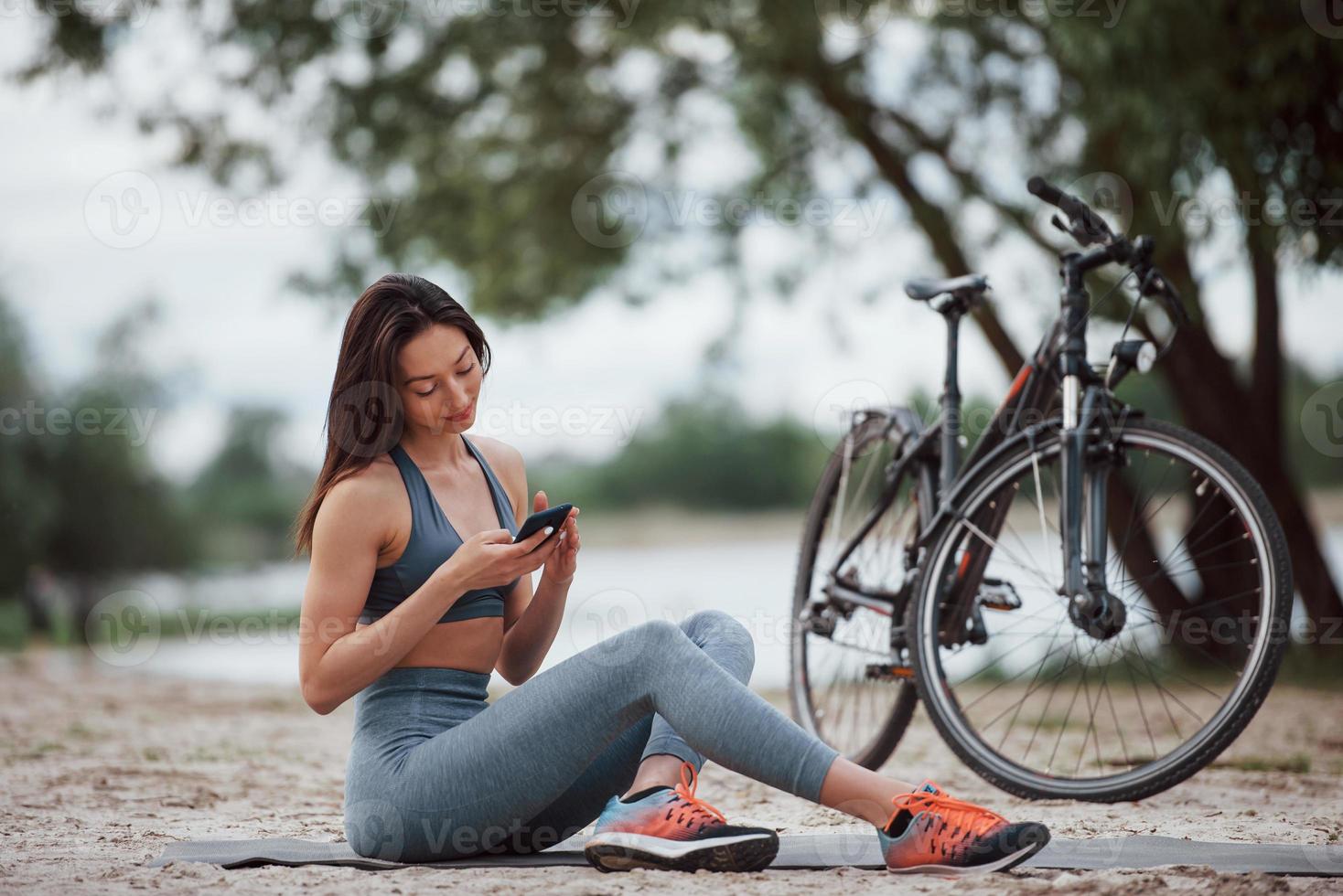 drahtlose Technologien. Radfahrerin mit guter Körperform, die tagsüber neben ihrem Fahrrad am Strand sitzt foto