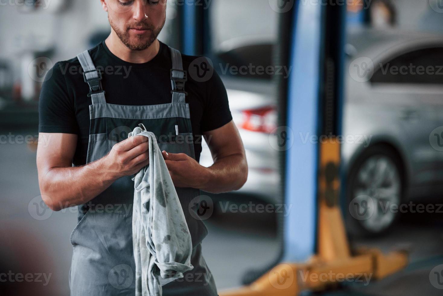 gewöhnlicher Arbeitstag. Mann in schwarzem Hemd und grauer Uniform steht in der Garage, nachdem er ein kaputtes Auto repariert hat foto